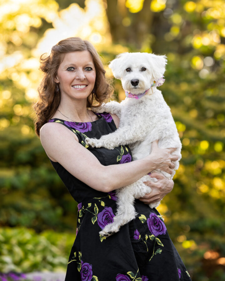 A woman in a black dress with purple flowers holds her white Schnauzer on her hip at Grand Ideas Gardens in Grand Rapids, MI.